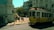 A vintage yellow tram is making its way through a street intersection, surrounded by historic European architecture. Several people are standing near the tram stop; some are taking photos, and others are simply observing. Tall buildings with ornate facades and a few trees can be seen in the background. The lighting suggests it is a sunny day, casting shadows on the ground.