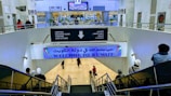 A large indoor area at an airport with signs in both Arabic and English, welcoming visitors to Kuwait. There are several people moving about, some on a downward escalator. The area is well-lit, with visible directions for arrivals, baggage claim, customs, and lost & found. Plant decorations and digital screens can also be seen.