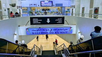 A large indoor area at an airport with signs in both Arabic and English, welcoming visitors to Kuwait. There are several people moving about, some on a downward escalator. The area is well-lit, with visible directions for arrivals, baggage claim, customs, and lost & found. Plant decorations and digital screens can also be seen.