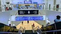 A large indoor area at an airport with signs in both Arabic and English, welcoming visitors to Kuwait. There are several people moving about, some on a downward escalator. The area is well-lit, with visible directions for arrivals, baggage claim, customs, and lost & found. Plant decorations and digital screens can also be seen.