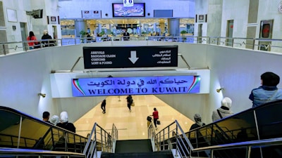 A large indoor area at an airport with signs in both Arabic and English, welcoming visitors to Kuwait. There are several people moving about, some on a downward escalator. The area is well-lit, with visible directions for arrivals, baggage claim, customs, and lost & found. Plant decorations and digital screens can also be seen.
