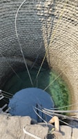 A deep stone well with clear water visible at the bottom. Multiple pipes and ropes are hanging into the well, suggesting it is actively used for drawing water. The circular stone walls are meticulously arranged, showing the traditional construction method.