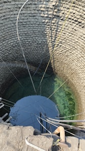 A deep stone well with clear water visible at the bottom. Multiple pipes and ropes are hanging into the well, suggesting it is actively used for drawing water. The circular stone walls are meticulously arranged, showing the traditional construction method.