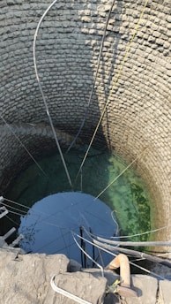 A deep stone well with clear water visible at the bottom. Multiple pipes and ropes are hanging into the well, suggesting it is actively used for drawing water. The circular stone walls are meticulously arranged, showing the traditional construction method.