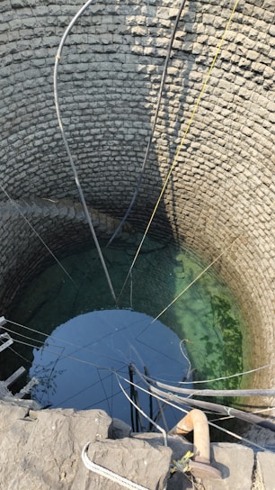 A deep stone well with clear water visible at the bottom. Multiple pipes and ropes are hanging into the well, suggesting it is actively used for drawing water. The circular stone walls are meticulously arranged, showing the traditional construction method.