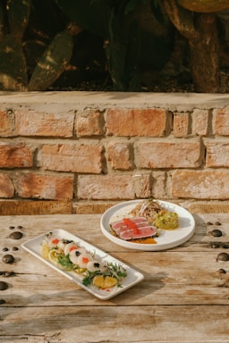 Two plates of elaborately styled gourmet dishes are set on a rustic wooden table against a brick wall. One plate contains seared tuna with a garnish of sprouts, sauce, and a side of mashed avocado topped with red fish roe. The other plate features a variety of sliced vegetables and citrus with elegantly arranged garnishes.