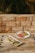 Two plates of elaborately styled gourmet dishes are set on a rustic wooden table against a brick wall. One plate contains seared tuna with a garnish of sprouts, sauce, and a side of mashed avocado topped with red fish roe. The other plate features a variety of sliced vegetables and citrus with elegantly arranged garnishes.