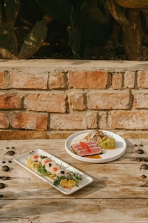 Two plates of elaborately styled gourmet dishes are set on a rustic wooden table against a brick wall. One plate contains seared tuna with a garnish of sprouts, sauce, and a side of mashed avocado topped with red fish roe. The other plate features a variety of sliced vegetables and citrus with elegantly arranged garnishes.
