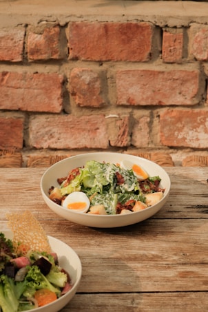 A rustic wooden table holds a bowl of salad featuring leafy greens, croutons, sliced boiled eggs, and bacon, topped with dressing. Another partial bowl of fresh mixed salad with various vegetables is also visible. The background features a brick wall, adding a warm, earthy backdrop to the scene.