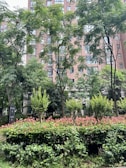 A lush, green landscape with a variety of trees and shrubs in front of a multi-story building. The building has numerous windows, and the foliage features various shades of green with some red-tipped leaves. A small sign is visible among the greenery.