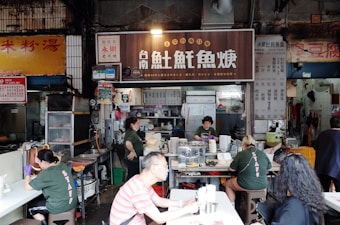 A bustling food stall with several people both working and dining. The main stall has a large sign with Asian characters, and inside, workers are preparing food. Customers sit at nearby tables. The setting appears to be a market or street food area with a variety of cooking utensils and dishes.