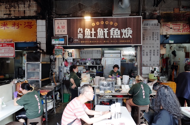 A bustling food stall with several people both working and dining. The main stall has a large sign with Asian characters, and inside, workers are preparing food. Customers sit at nearby tables. The setting appears to be a market or street food area with a variety of cooking utensils and dishes.