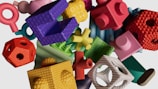 A collection of multicolored, textured geometric shapes are scattered on a light background. The objects vary in form, including cubes, spheres, rings, and complex shapes, each with distinct surface textures such as bumps and grooves. The arrangement appears random, creating a visually engaging and playful composition.