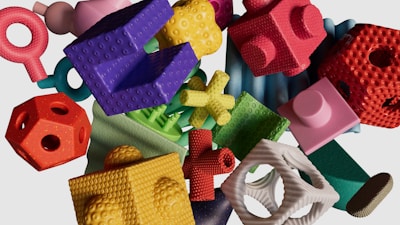 A collection of multicolored, textured geometric shapes are scattered on a light background. The objects vary in form, including cubes, spheres, rings, and complex shapes, each with distinct surface textures such as bumps and grooves. The arrangement appears random, creating a visually engaging and playful composition.