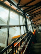 A modern indoor staircase with black metal railings leading upwards. Large glass windows on the left allow daylight to stream in, casting reflections on the surfaces. The structure features steel beams and an industrial design with orange accents visible inside and through the windows.