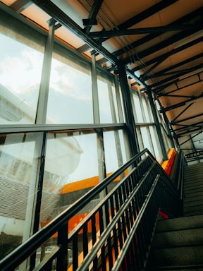 A modern indoor staircase with black metal railings leading upwards. Large glass windows on the left allow daylight to stream in, casting reflections on the surfaces. The structure features steel beams and an industrial design with orange accents visible inside and through the windows.