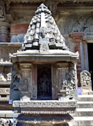 Intricate stone carvings adorn an ancient architectural structure. The central focus is a small shrine embedded with detailed sculptures of deities and figures, showcasing expertise in stone artistry. The structure features embellishments and motifs typical of traditional temple design.