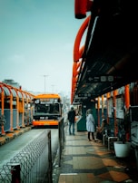 A modern, orange bus labeled 'Bus Listrik' is parked at a bus stop in an urban area. The bus stop features bright orange and gray accents, with several people waiting, including a person sitting and another standing and conversing. The area appears organized and bustling, with an overcast sky visible in the background.