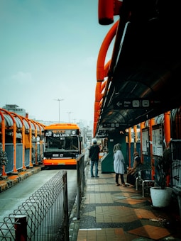 A modern, orange bus labeled 'Bus Listrik' is parked at a bus stop in an urban area. The bus stop features bright orange and gray accents, with several people waiting, including a person sitting and another standing and conversing. The area appears organized and bustling, with an overcast sky visible in the background.