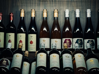 A collection of various wine bottles is neatly arranged on a wooden shelf. The labels on the bottles display different types and flavors, including Shiraz, Cabernet, Sauvignon Blanc, and Pomegranate. Some bottles have gold or red caps, adding a variety of colors to the arrangement. The background is a wooden texture, enhancing the rustic and sophisticated atmosphere.