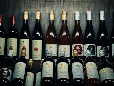 A collection of various wine bottles is neatly arranged on a wooden shelf. The labels on the bottles display different types and flavors, including Shiraz, Cabernet, Sauvignon Blanc, and Pomegranate. Some bottles have gold or red caps, adding a variety of colors to the arrangement. The background is a wooden texture, enhancing the rustic and sophisticated atmosphere.