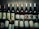 A collection of various wine bottles is neatly arranged on a wooden shelf. The labels on the bottles display different types and flavors, including Shiraz, Cabernet, Sauvignon Blanc, and Pomegranate. Some bottles have gold or red caps, adding a variety of colors to the arrangement. The background is a wooden texture, enhancing the rustic and sophisticated atmosphere.