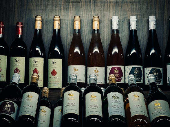 A collection of various wine bottles is neatly arranged on a wooden shelf. The labels on the bottles display different types and flavors, including Shiraz, Cabernet, Sauvignon Blanc, and Pomegranate. Some bottles have gold or red caps, adding a variety of colors to the arrangement. The background is a wooden texture, enhancing the rustic and sophisticated atmosphere.