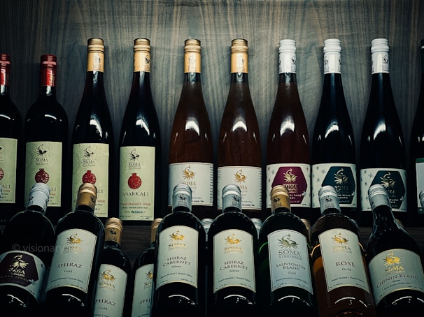 A collection of various wine bottles is neatly arranged on a wooden shelf. The labels on the bottles display different types and flavors, including Shiraz, Cabernet, Sauvignon Blanc, and Pomegranate. Some bottles have gold or red caps, adding a variety of colors to the arrangement. The background is a wooden texture, enhancing the rustic and sophisticated atmosphere.