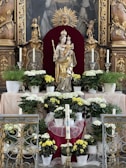 A religious altar adorned with a statue of a crowned female figure holding a child, surrounded by white and yellow flowers in pots. The background features ornate carvings and a decorative golden sunburst. Candles are placed symmetrically on either side of the statue, along with intricate metalwork in the foreground.