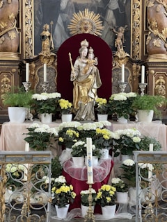 A religious altar adorned with a statue of a crowned female figure holding a child, surrounded by white and yellow flowers in pots. The background features ornate carvings and a decorative golden sunburst. Candles are placed symmetrically on either side of the statue, along with intricate metalwork in the foreground.