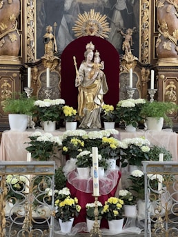 A religious altar adorned with a statue of a crowned female figure holding a child, surrounded by white and yellow flowers in pots. The background features ornate carvings and a decorative golden sunburst. Candles are placed symmetrically on either side of the statue, along with intricate metalwork in the foreground.