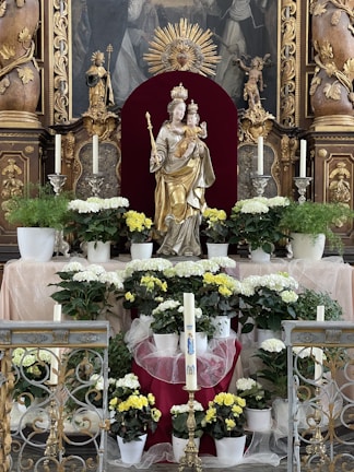 A religious altar adorned with a statue of a crowned female figure holding a child, surrounded by white and yellow flowers in pots. The background features ornate carvings and a decorative golden sunburst. Candles are placed symmetrically on either side of the statue, along with intricate metalwork in the foreground.