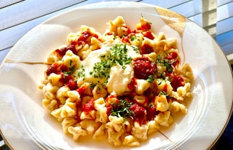A plate of pasta featuring a white sauce topped with tomato sauce and garnished with chopped herbs, presented on an elegant white and gold-rimmed plate.