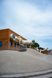 A brick structure features an interesting architectural design with multiple large, white tubular slides protruding from the building. The building is situated behind a sloping, sandy-colored landscape. Metal railings curve along the contour of the slope, and several windows are visible on the side of the structure. The sky is mostly clear, creating a spacious and open atmosphere.