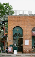 A brick building with a shop front featuring a green arched door and an ampersand symbol above it. A spiral staircase is visible on the left side next to a tree. There are patio tables and chairs set out in front of the shop. Signs with pet-friendly messages are displayed near the entrance.