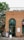 A brick building with a shop front featuring a green arched door and an ampersand symbol above it. A spiral staircase is visible on the left side next to a tree. There are patio tables and chairs set out in front of the shop. Signs with pet-friendly messages are displayed near the entrance.