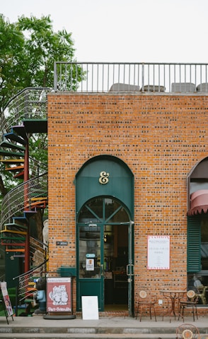 A brick building with a shop front featuring a green arched door and an ampersand symbol above it. A spiral staircase is visible on the left side next to a tree. There are patio tables and chairs set out in front of the shop. Signs with pet-friendly messages are displayed near the entrance.