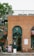 A brick building with a shop front featuring a green arched door and an ampersand symbol above it. A spiral staircase is visible on the left side next to a tree. There are patio tables and chairs set out in front of the shop. Signs with pet-friendly messages are displayed near the entrance.