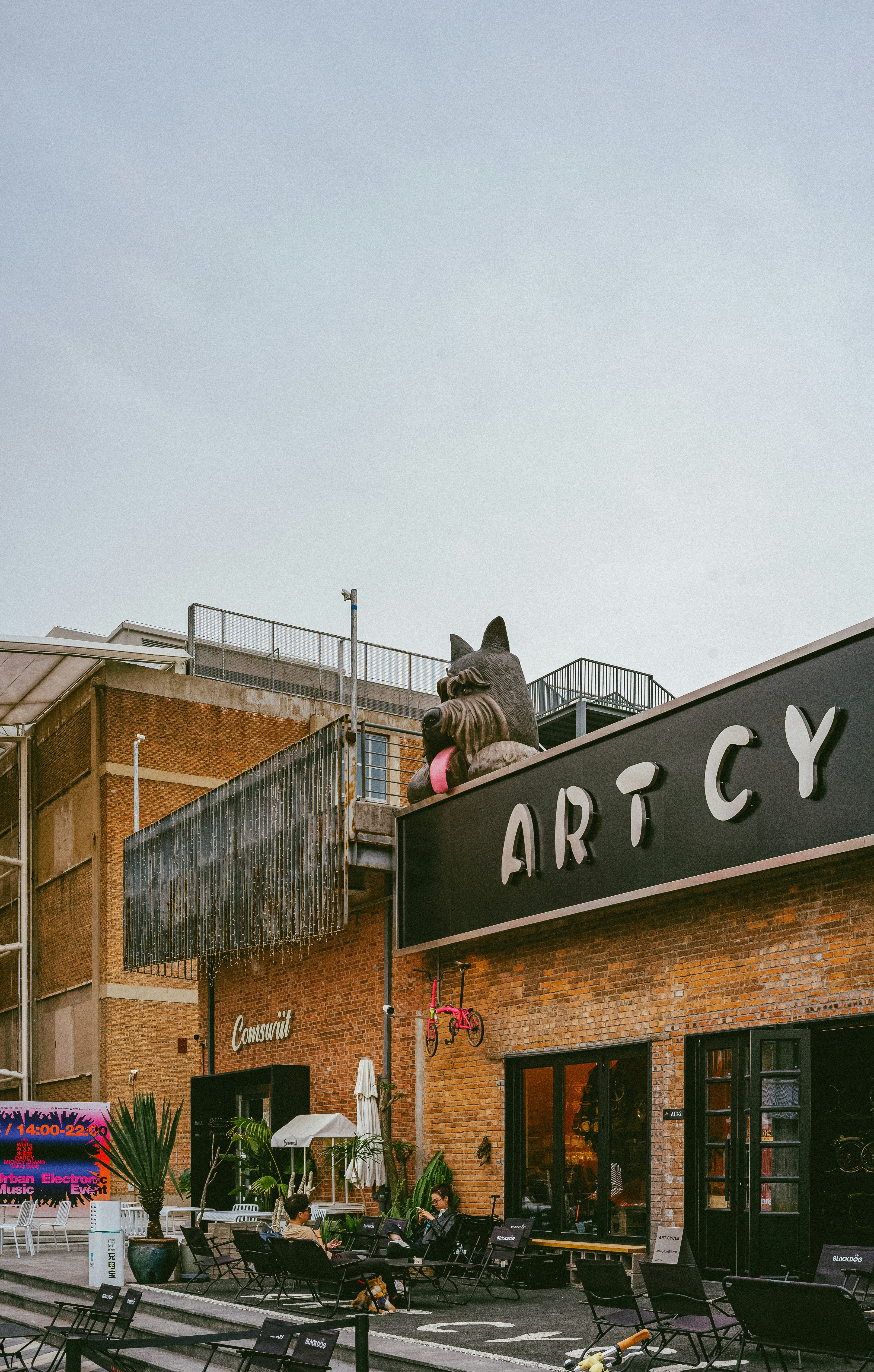 A large outdoor space featuring a modern brick building with the word 'ARTCY' prominently displayed. A giant sculpture of a dog with sunglasses is positioned on top of the building. Several people are seated at tables on the patio in front of the building, which is decorated with plants and signage. The area has a casual, artistic vibe.