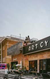 A large outdoor space featuring a modern brick building with the word 'ARTCY' prominently displayed. A giant sculpture of a dog with sunglasses is positioned on top of the building. Several people are seated at tables on the patio in front of the building, which is decorated with plants and signage. The area has a casual, artistic vibe.