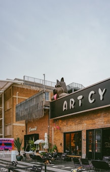 A large outdoor space featuring a modern brick building with the word 'ARTCY' prominently displayed. A giant sculpture of a dog with sunglasses is positioned on top of the building. Several people are seated at tables on the patio in front of the building, which is decorated with plants and signage. The area has a casual, artistic vibe.