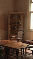 A vintage room featuring a wooden table with a weathered surface. An ornate cabinet with glass doors stands against the wall, displaying various decorative items such as porcelain dishes and small vases. A couple of classic wooden chairs with upholstered seats are placed around the table. The room is softly lit by natural light streaming through a window with white frames, casting an inviting and nostalgic atmosphere.