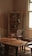 A vintage room featuring a wooden table with a weathered surface. An ornate cabinet with glass doors stands against the wall, displaying various decorative items such as porcelain dishes and small vases. A couple of classic wooden chairs with upholstered seats are placed around the table. The room is softly lit by natural light streaming through a window with white frames, casting an inviting and nostalgic atmosphere.