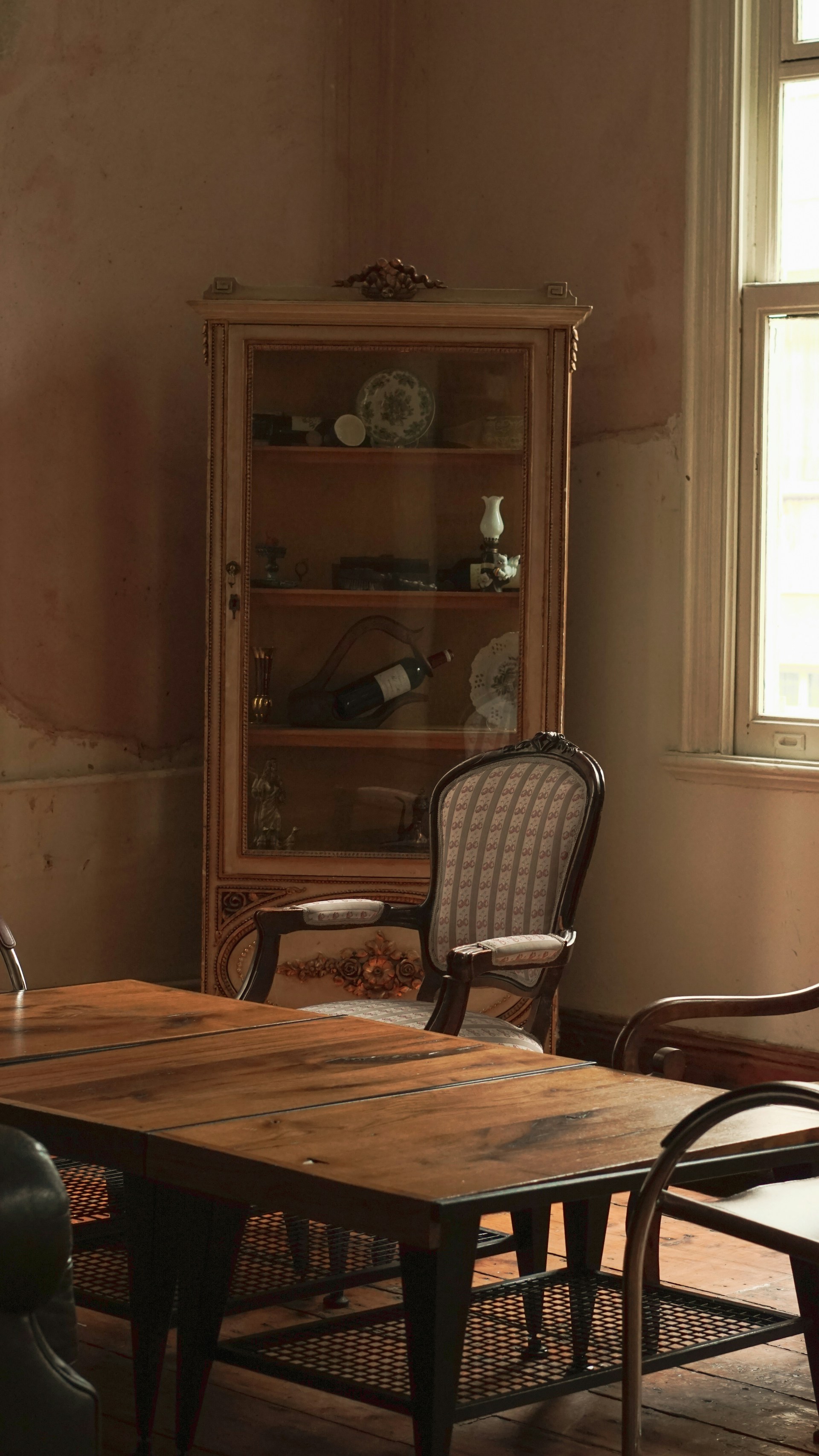 A vintage room featuring a wooden table with a weathered surface. An ornate cabinet with glass doors stands against the wall, displaying various decorative items such as porcelain dishes and small vases. A couple of classic wooden chairs with upholstered seats are placed around the table. The room is softly lit by natural light streaming through a window with white frames, casting an inviting and nostalgic atmosphere.