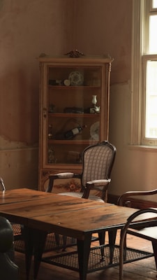 A vintage room featuring a wooden table with a weathered surface. An ornate cabinet with glass doors stands against the wall, displaying various decorative items such as porcelain dishes and small vases. A couple of classic wooden chairs with upholstered seats are placed around the table. The room is softly lit by natural light streaming through a window with white frames, casting an inviting and nostalgic atmosphere.