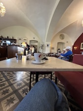 A cozy cafe setting with arched ceilings and elegant lighting. A table in the foreground holds a white coffee cup and saucer with a red logo. In the background, several people are seated wearing hats, while others stand at the counter. The room is decorated with framed pictures and has patterned floor tiles.