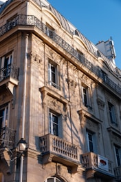 A classic European-style building facade with ornate details and wrought iron balconies. The sunlight casts intricate shadows across the cream-colored stone exterior, highlighting its architectural features.
