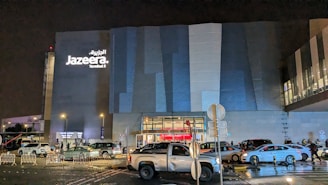 A modern building facade with the Jazeera Terminal 5 signage illuminated at night. The scene is bustling with several vehicles and people visible in the foreground, likely in a parking or drop-off area. The building features a sleek design with horizontal paneling, and there are street lamps providing additional lighting.