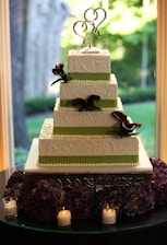 A beautifully decorated four-tiered wedding cake with intricate white icing patterns and green ribbons. It features a metal heart-shaped cake topper. The base is surrounded by purple flowers and lit candles, creating an elegant and romantic atmosphere with a lush garden visible through the window behind.