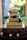 A beautifully decorated four-tiered wedding cake with intricate white icing patterns and green ribbons. It features a metal heart-shaped cake topper. The base is surrounded by purple flowers and lit candles, creating an elegant and romantic atmosphere with a lush garden visible through the window behind.